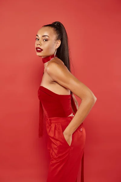 Beautiful african american model in red strapless top and pants posing with hand in pocket — Stock Photo