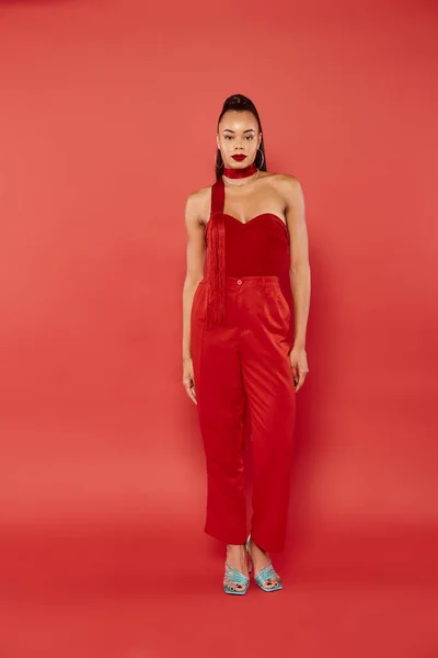 Longitud completa de atractivo modelo afroamericano en top sin tirantes y pantalones posando sobre fondo rojo - foto de stock