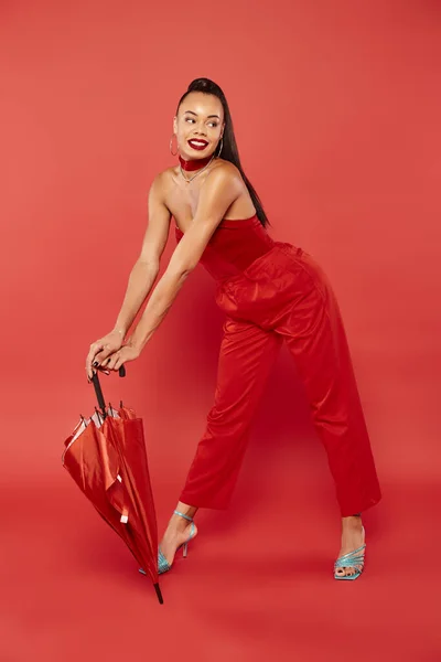 Piena lunghezza di allegro modello afroamericano in rosso senza spalline top e pantaloni in posa con ombrello — Foto stock