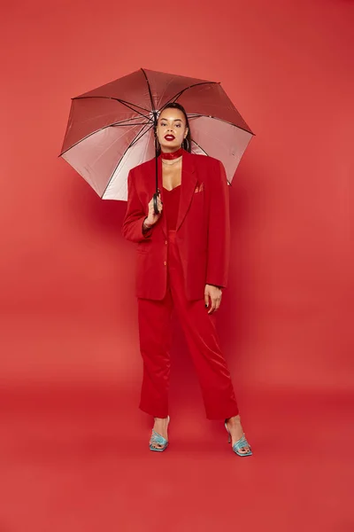 Full length, brunette african american model in red blazer and pants standing under umbrella — Stock Photo