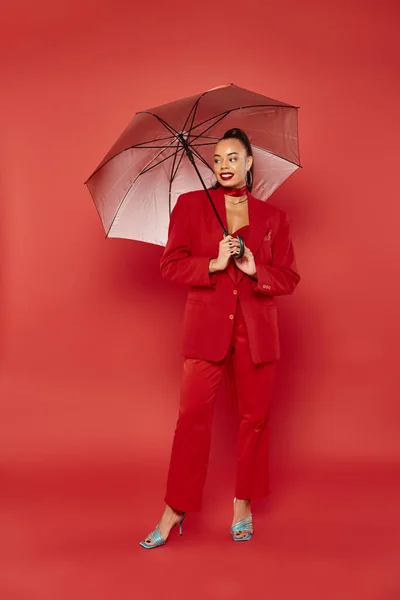 Larga duración, modelo americano africano alegre en chaqueta roja y pantalones de pie bajo paraguas - foto de stock
