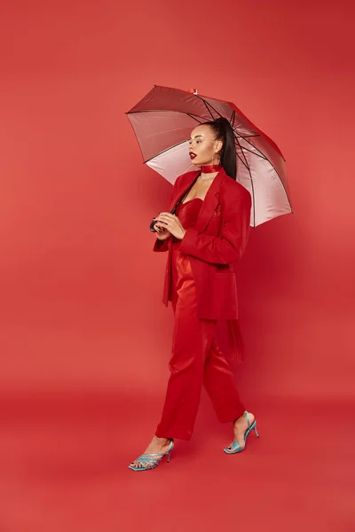 Bonita africana americana mujer en chaqueta y pantalones caminando bajo paraguas en rojo telón de fondo - foto de stock