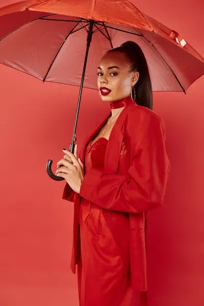 Pretty african american woman in jacket and pants standing under umbrella on red backdrop — Stock Photo