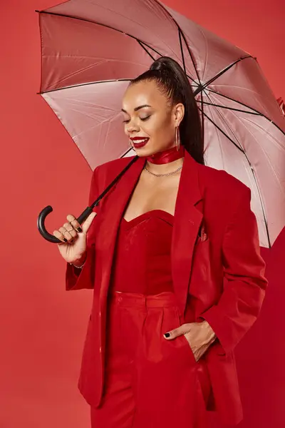 Sonriente mujer afroamericana en blazer y pantalones de pie bajo paraguas sobre fondo rojo - foto de stock