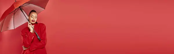 Attractive young african american woman in blazer and pants standing under umbrella on red, banner — Stock Photo
