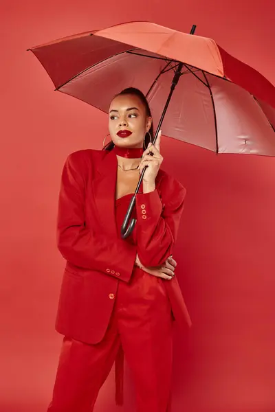 Attractive young african american woman in jacket and pants standing under umbrella on red backdrop — Stock Photo