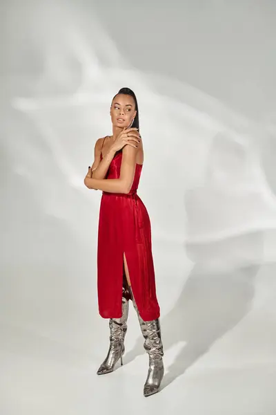 Pleine longueur de femme afro-américaine en robe rouge et bottes argentées debout sur fond gris — Photo de stock