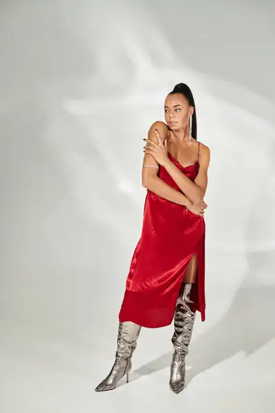 Pleine longueur de femme brune afro-américaine en robe rouge et bottes argentées sur fond gris — Photo de stock