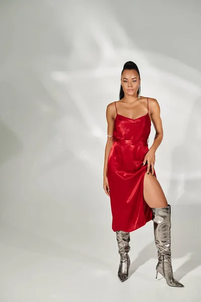 Полная длина соблазнительной африканской американки в красном платье и серебряных сапогах на сером фоне — стоковое фото