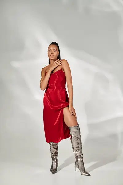 Longitud completa de la glamorosa mujer afroamericana en vestido rojo y botas de plata sobre fondo gris - foto de stock