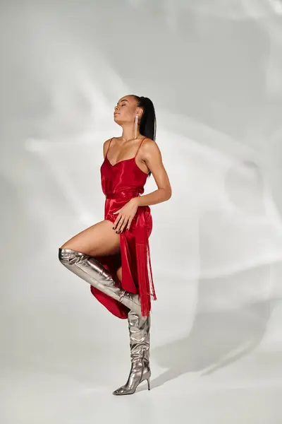 Longitud completa, atractiva mujer afroamericana en vestido rojo y botas de plata posando sobre fondo gris - foto de stock