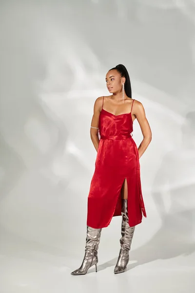 Longitud completa, hermosa mujer afroamericana en vestido rojo y botas de plata posando sobre fondo gris - foto de stock