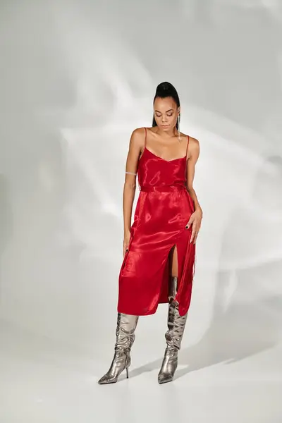 Полная длина, африканская американка в красном платье миди и серебряных сапогах, позирующих на сером фоне — стоковое фото