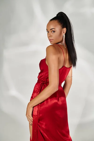 Bonita mulher americana africana em vestido vermelho olhando para a câmera e posando em fundo cinza espelhado — Fotografia de Stock