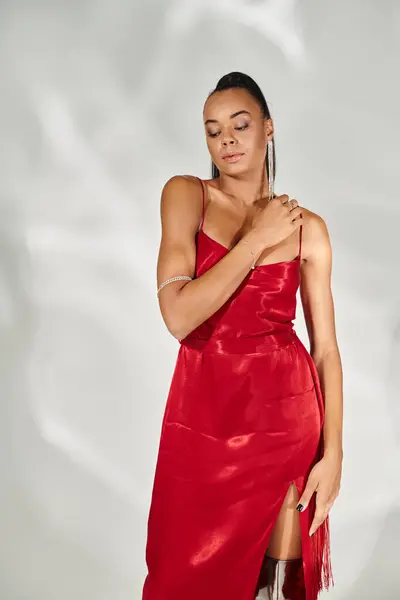 Mujer afroamericana sensual en vestido rojo mirando hacia otro lado y posando sobre fondo gris espejado - foto de stock