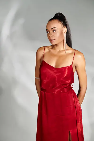 Hermosa mujer afroamericana en vestido rojo mirando hacia otro lado y posando sobre fondo gris espejo - foto de stock