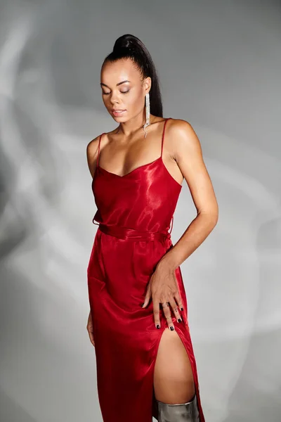 Elegante mujer afroamericana en vestido rojo mirando hacia abajo y posando sobre fondo gris espejado - foto de stock