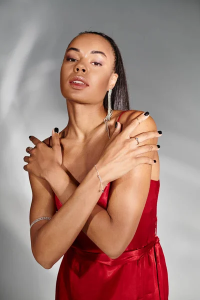 Glamorosa mujer afroamericana en vestido rojo cruzando brazos al posar sobre fondo gris espejado - foto de stock