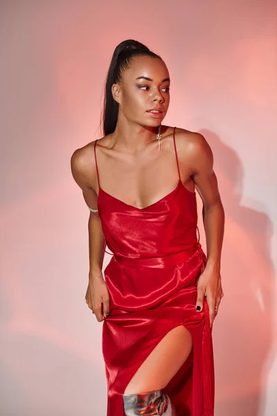 Atractiva mujer afroamericana en vestido rojo mirando hacia otro lado sobre fondo gris con iluminación - foto de stock