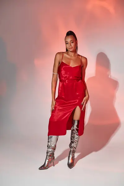 Jolie femme afro-américaine en robe rouge midi et bottes argentées sur fond gris avec éclairage — Photo de stock