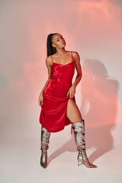 Full length, african american woman in red midi dress and silver boots posing on grey with lighting — Stock Photo