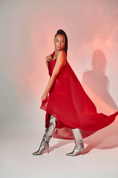 Afro-américaine en robe rouge et châle marchant en bottes argentées sur gris avec éclairage — Photo de stock