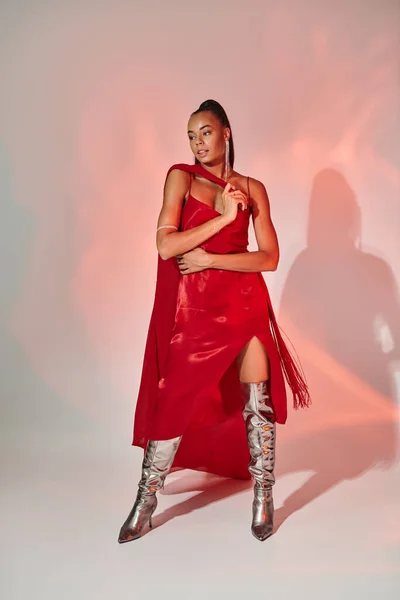 Pretty african american woman in red dress with shawl standing in silver boots on grey with lighting — Stock Photo