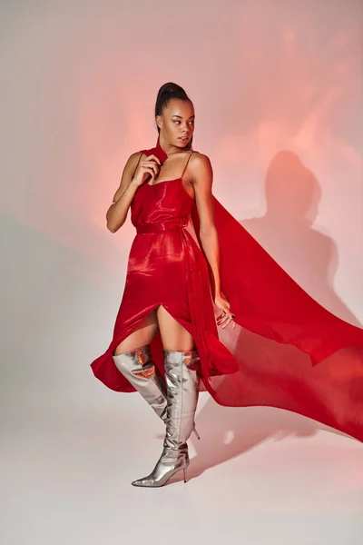 Joven mujer afroamericana en vestido rojo con chal de pie en botas de plata en gris con iluminación - foto de stock