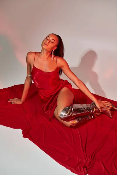 Longitud completa, mujer afroamericana en vestido midi rojo y botas de plata sentado en gris con iluminación - foto de stock