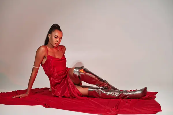 Attractive african american woman in red midi dress and silver boots sitting on grey with lighting — Stock Photo