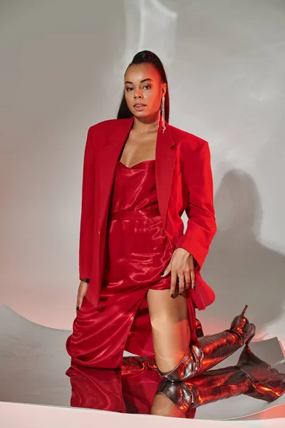 Junge Afroamerikanerin in rotem Kleid, Blazer und silbernen Stiefeln posiert auf verspiegelter Oberfläche — Stockfoto