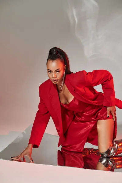 Joven modelo afroamericano en vestido rojo, chaqueta y botas plateadas posando sobre superficie espejada - foto de stock