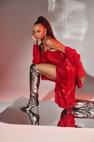 Atractivo modelo afroamericano en blazer rojo, vestido y botas de plata posando en superficie espejada - foto de stock
