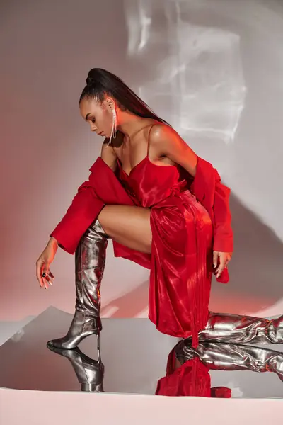 Morena modelo afroamericano en blazer rojo, vestido y botas plateadas posando en superficie espejada - foto de stock