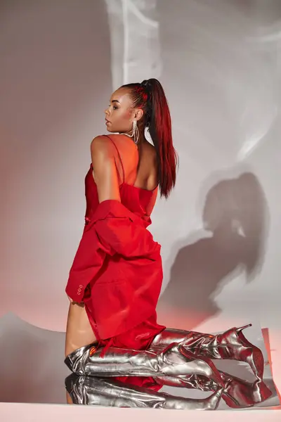 Encantador modelo afroamericano en blazer rojo, vestido y botas de plata posando en superficie espejada - foto de stock