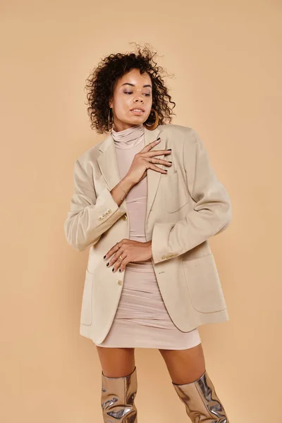 Morena mujer afroamericana con el pelo rizado posando en chaqueta otoñal y vestido en beige - foto de stock