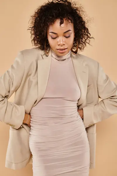 Slim african american woman with brunette curly hair posing in autumnal blazer and dress on beige — Stock Photo