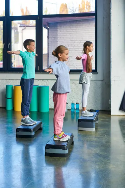Three adolescent girls and boy in sportswear exercising on fitness stepper with arms asides, sport — Stock Photo