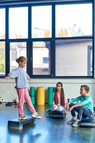 Bonita chica de pie en una pierna en fitness stepper con otros niños mirándola, deporte infantil - foto de stock