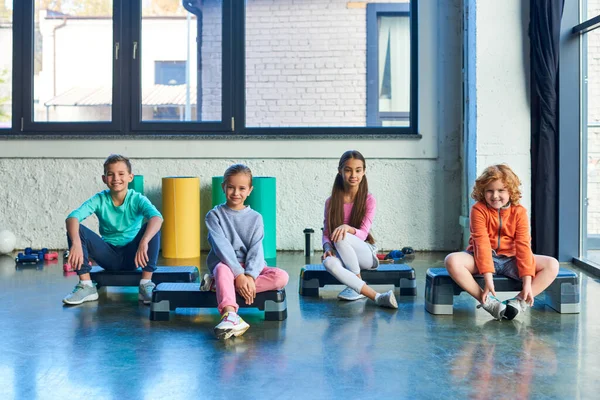 Cuatro niños y niñas preadolescentes sentados en pasos de fitness y sonriendo a la cámara, deporte infantil - foto de stock