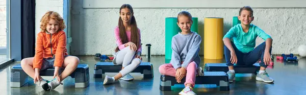 Cuatro lindos niños pequeños sentados en pasos de fitness y sonriendo a la cámara, deporte infantil, pancarta - foto de stock