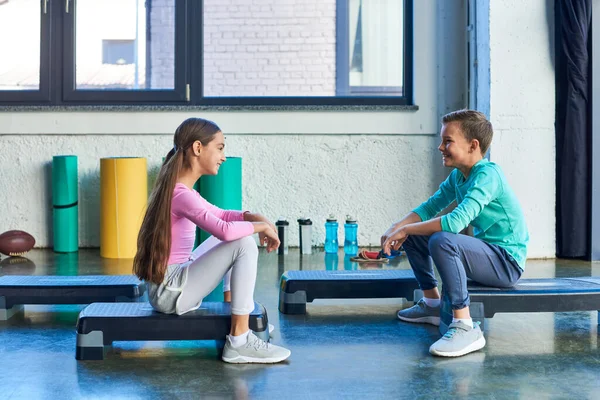 Alegre chico y chica sentado en fitness steppers y sonriendo felizmente el uno al otro, deporte infantil - foto de stock