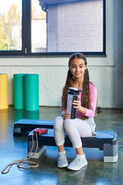 Vertikale Aufnahme des kleinen Smiley-Mädchens auf Fitness-Stepper sitzend mit Wasserflasche, Kindersport — Stockfoto