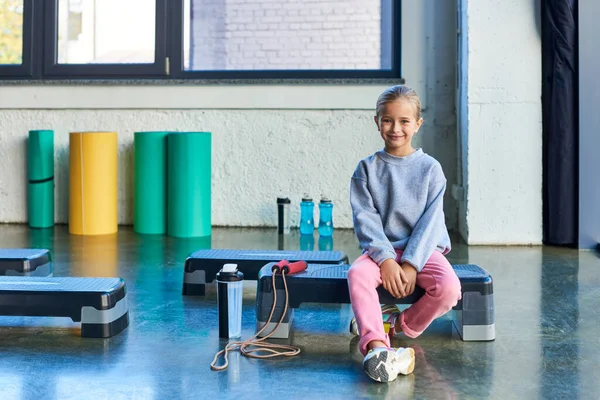 Весела блондинка маленька дівчинка сидить на фітнес-стрічці в спортивному одязі, посміхаючись на камеру, дитячий спорт — стокове фото