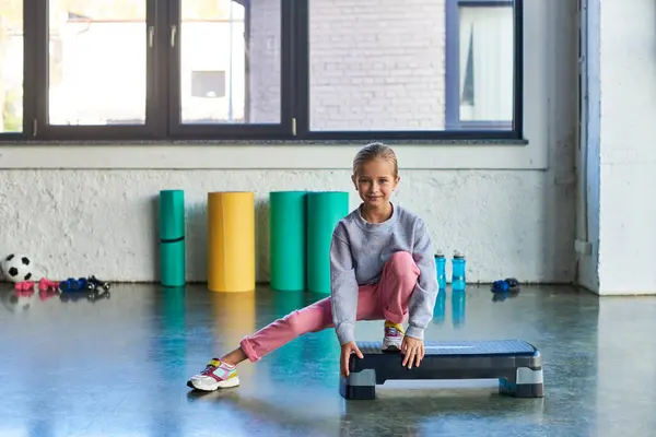 Kleines hübsches Mädchen in Sportbekleidung streckt Bein auf Fitness-Stepper schaut in die Kamera, Kindersport — Stockfoto