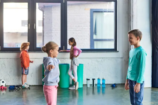 Blondes Mädchen und kleiner Junge, die in der Turnhalle trainieren, während ihre Freunde über die Kulisse, den Kindersport sprechen — Stockfoto