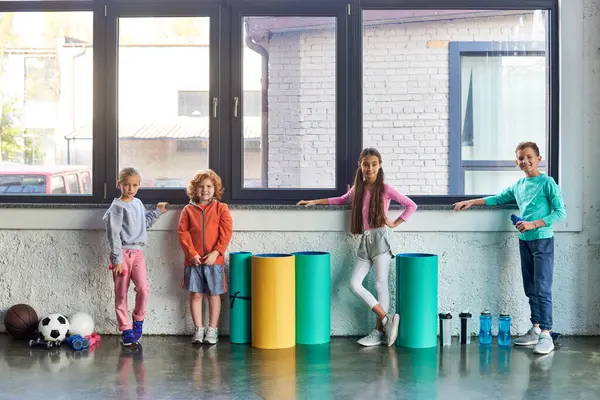 Cuatro niños y niñas lindos preadolescentes en ropa deportiva posando por la ventana en el gimnasio, deporte infantil - foto de stock