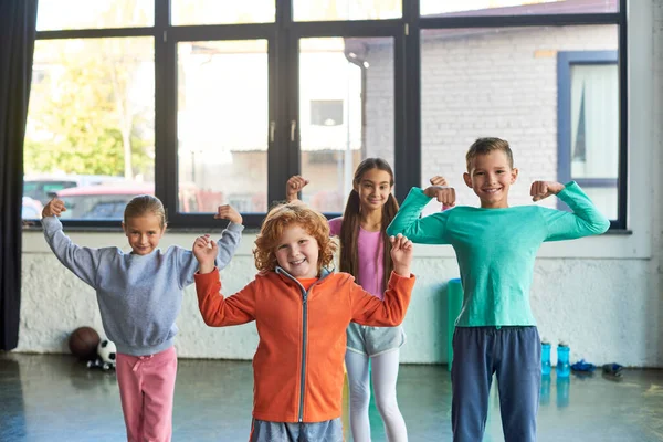 Cuatro niños pequeños y bonitos en ropa deportiva jugando músculos y sonriendo a la cámara, deporte infantil - foto de stock