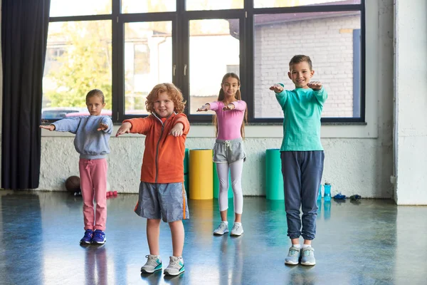 Quatro crianças pré-adolescentes alegres em roupas esportivas alongamento e sorrindo para a câmera, esporte infantil — Fotografia de Stock