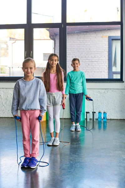 Tiro vertical de las niñas bonitas y el niño con cuerdas de salto en las manos sonriendo a la cámara, deporte infantil - foto de stock